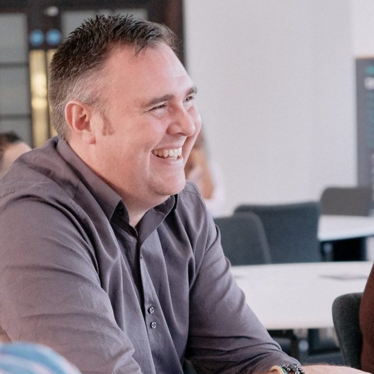 Smiling man in a grey shirt, seated at a table in a modern office setting.
